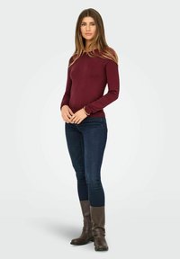 Bordeaux lange mouwen aansluitende trui met ronde hals, gecombineerd met donkerblauwe skinny jeans en kniehoge bruine leren laarzen.
