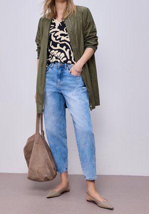 Kobieta w jasnoniebieskich jeansach, bluzce w zwierzęcy wzór, oliwkowej kurtce, beżowych balerinach, trzymająca dużą brązową zamszową torebkę.