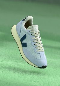 Sapatilha atlética azul clara com materiais em malha e camurça, detalhes em azul marinho, atacadores bege e sola em cor creme. Design arredondado, com branding minimalista.