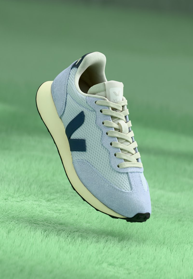 Sapatilha atlética azul clara com materiais em malha e camurça, detalhes em azul marinho, atacadores bege e sola em cor creme. Design arredondado, com branding minimalista.