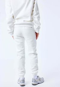 Sudadera blanca con puños acanalados, combinada con joggers blancos a juego que cuentan con un bolsillo trasero, llevada con zapatillas deportivas plateadas y blancas.