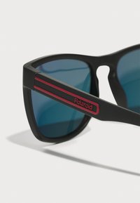 Ochelari de soare negri cu lentile albastre, având finisaj mat, dungi accent roșii pe tâmple și un logo Polaroid proeminent.
