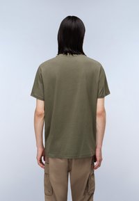 Camiseta de algodón suave de color verde oliva y manga corta. Presenta un escote redondeado y un corte relajado, combinada con pantalones cargo de color marrón claro.