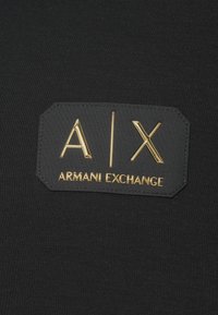 Crni materijal s ušivenim crnim flasterom na kojem se nalazi zlatni metalik natpis "A | X" i "ARMANI EXCHANGE".