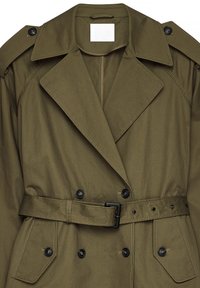 Olive krāsas tranšejas mantu apvalks, izgatavots no kokvilnas un sintētisko šķiedru apvienojuma. Tam ir divkāršs priekšpuse, liels apkakle un josta ar pogu akcentiem.