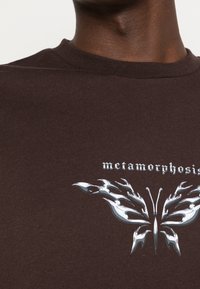 T-shirt en coton marron avec un graphique de papillon métallisé et des accents en forme de flamme, avec le mot "métamorphose" imprimé au-dessus en texte blanc.
