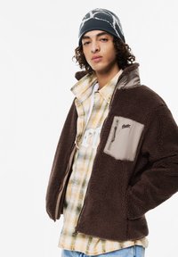 Brązowa polarowa kurtka z szarym panelem i kieszenią na zamek, noszona na koszuli w kratkę. Na głowie szara i biała czapka z wzorem.