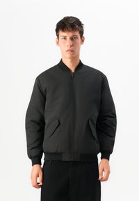 Schwarze Bomberjacke mit durchgehendem Reißverschluss, geripptem Kragen und Bündchen. Verfügt über seitliche Taschen und eine glatte, matte Textur.