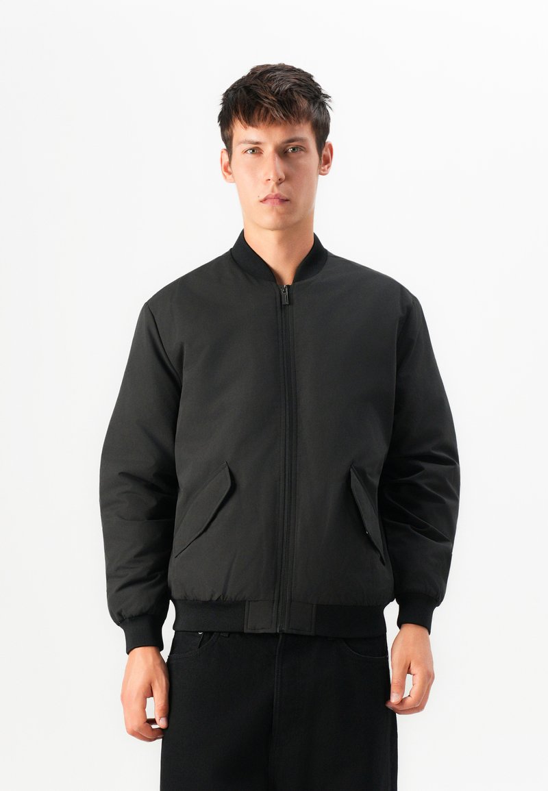 Schwarze Bomberjacke mit durchgehendem Reißverschluss, geripptem Kragen und Bündchen. Verfügt über seitliche Taschen und eine glatte, matte Textur.