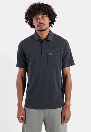 Homme aux cheveux bouclés portant un polo gris foncé avec une poche poitrine et un pantalon gris clair, debout devant un fond blanc.