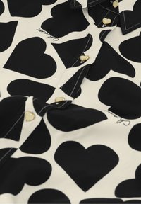 Zwart-wit shirt met een hartvormig patroon, voorzien van gouden knopen in de vorm van hartjes en een omgeslagen kraag. Zachte stof met een gladde textuur.