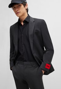 Kolkgray kostym med slim fit, kombinerad med en svart skjorta med knappar. Anmärkningsvärd röd märkespåse på ärmen. Svart keps fulländar looken.