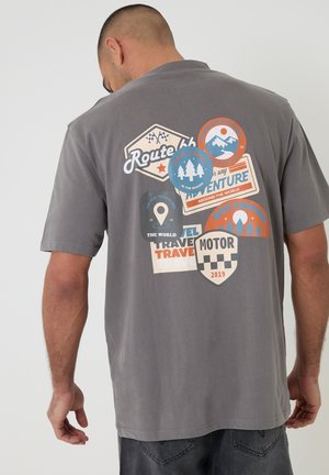 Mand iført grå t-shirt med farverige patches med temaer om rejser og eventyr på ryggen, herunder Route 66 og campingdesigns.