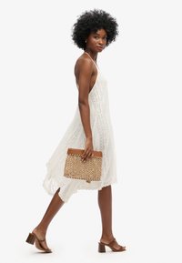 Weißes Spitzenkleid mit lockerer Passform, knielangem Saum und dünnen Trägern. Das Model hält eine tanfarbene gewebte Clutch. Braune Blockabsatzsandalen.