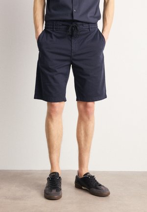 Pantaloni corti in cotone blu navy con vita elastica e cordoncino, dotati di due tasche laterali e un taglio dritto. Abbinati a sneakers nere.
