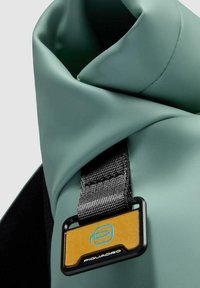 Material de bolsa impermeable verde con un borde doblado, acentuado por una correa negra y una etiqueta de marca amarilla y azul. Textura suave, diseño práctico.