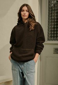 Brązowy polarowy sweatshirt z kapturem i przednią kieszenią, w połączeniu z jasnoniebieskimi dżinsami o luźnym kroju i szerokich nogawkach.