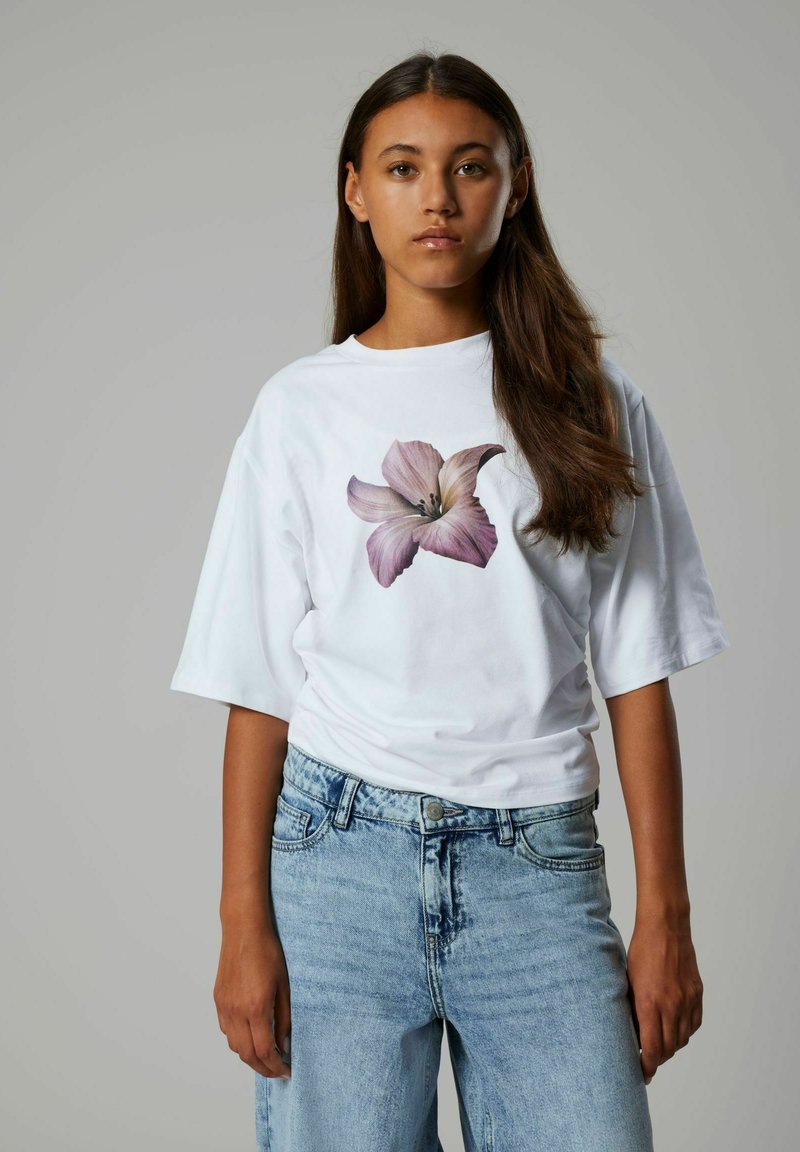 Camiseta blanca con un gran gráfico de lirio rosa, corte holgado, cuello redondo, combinada con jeans de mezclilla azul claro, destacando un diseño informal.