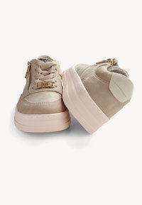 Baskets en daim beige avec une finition texturée, dotées de détails en doré, d'accents blancs et d'une semelle rose épaisse. Design à lacets avec zip sur le côté.
