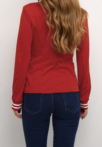 Rød langærmet top med tætsiddende pasform og diskrete sømdetaljer. Manchetterne har hvide striber. Bæres med mørkeblå jeans med glat tekstur.