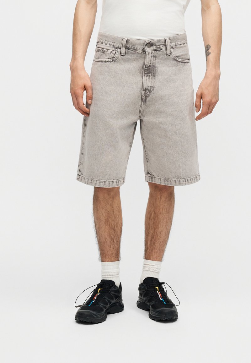 Hombre con pantalones cortos de mezclilla gris claro, calcetines blancos y zapatillas negras, de pie sobre un fondo claro y liso.