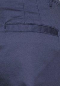 Close-up van een donkerblauwe stoffen zak met zichtbare stiksels op een kledingstuk, dat een horizontale openingen heeft.