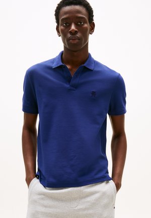 Junger Mann steht mit Händen in den Taschen, trägt ein blaues kurzärmeliges Poloshirt und hellgraue Hosen vor einem schlichten Hintergrund.