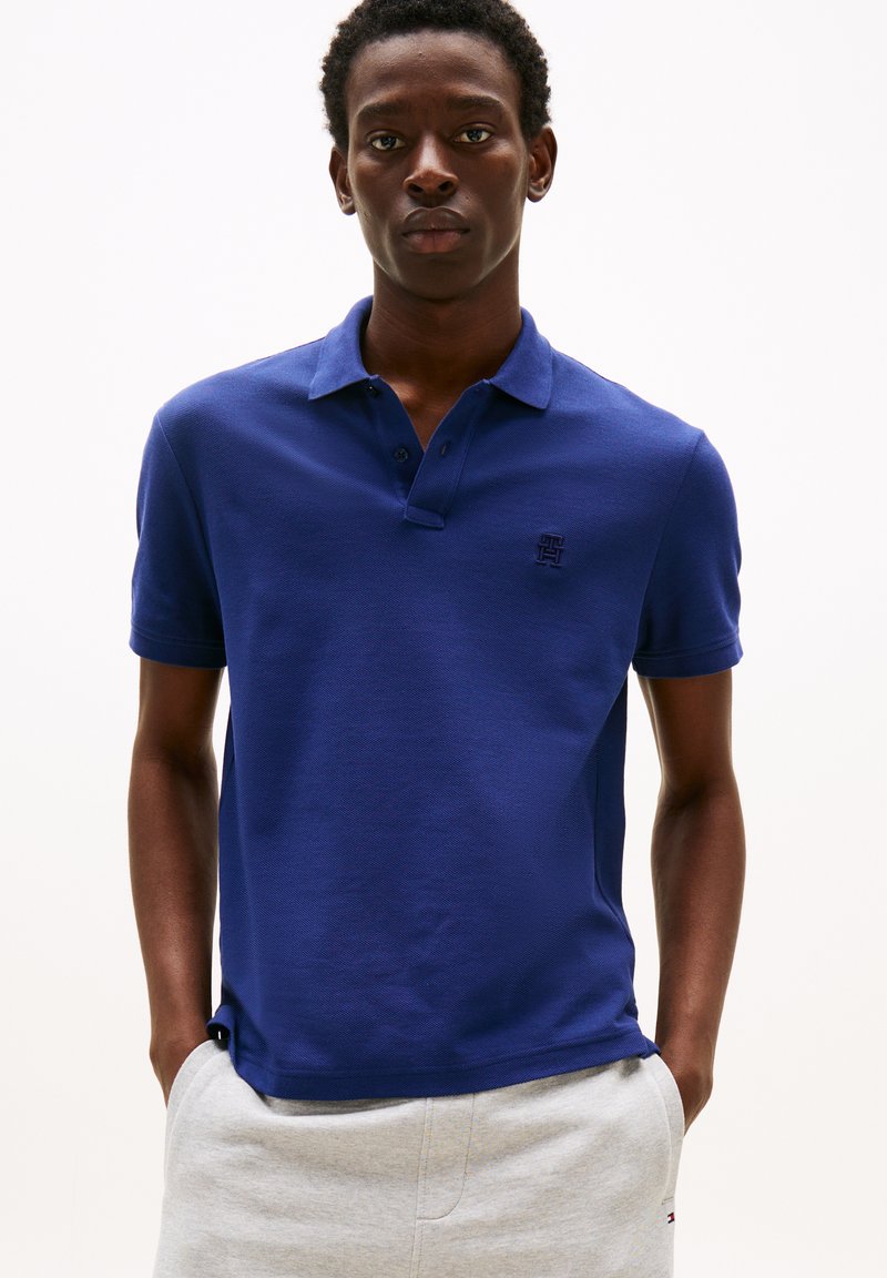 Junger Mann steht mit Händen in den Taschen, trägt ein blaues kurzärmeliges Poloshirt und hellgraue Hosen vor einem schlichten Hintergrund.