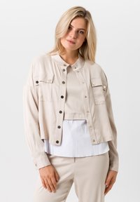 Beige cropped jasje met drukknoopsluiting, twee borstzakken en een mandarin kraag. Lichtgewicht stof met een gladde textuur.