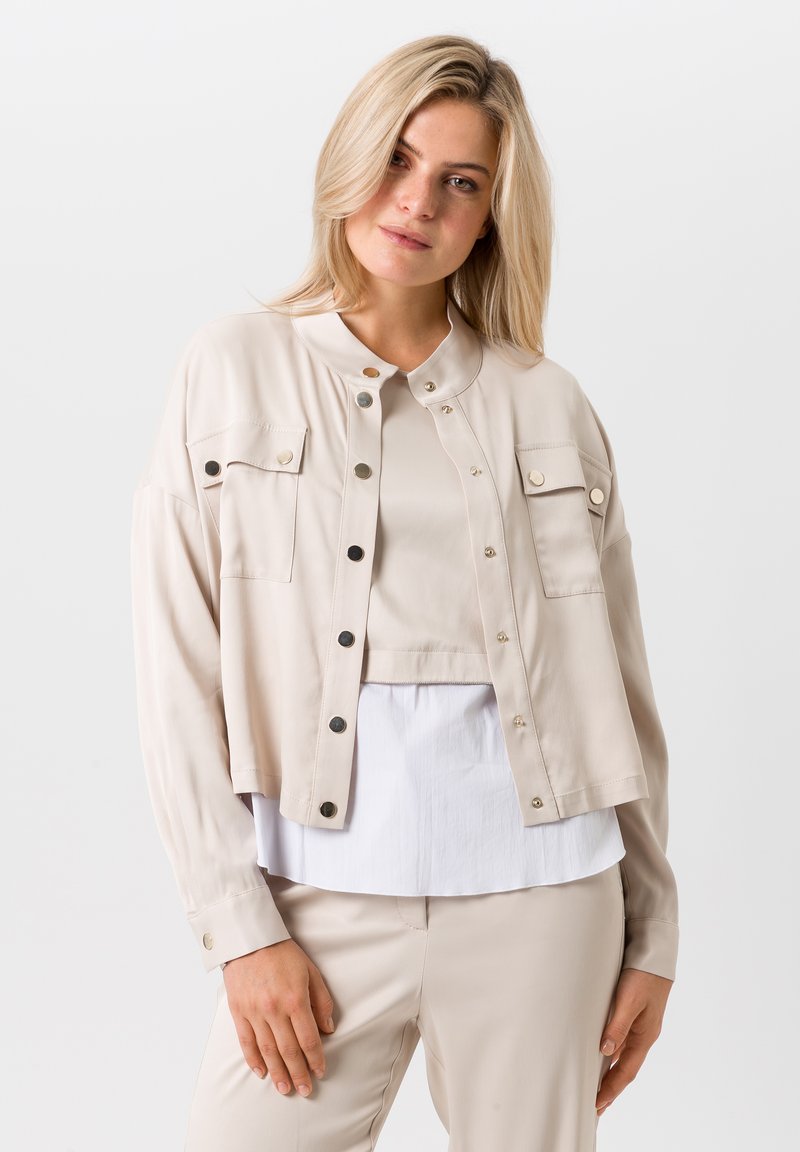 Beige cropped jasje met drukknoopsluiting, twee borstzakken en een mandarin kraag. Lichtgewicht stof met een gladde textuur.