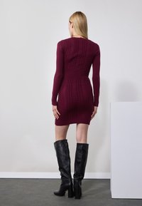 Robe en maille bordeaux avec des manches longues et un motif en relief. Associée à des bottes en cuir noires montant jusqu'aux genoux et à une silhouette ajustée.