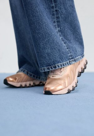 Personne portant des baskets en or rose métallique et un jean large en denim bleu, debout sur un sol bleu clair.