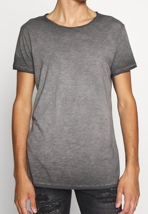 Homme portant un t-shirt gris délavé à manches courtes et col rond, ainsi qu'un jean noir déchiré, debout devant un fond clair uni.