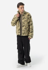 Fleecejakke i khaki med camouflage mønster, med høj krave, fuld lynlås og en tekstureret finish. Sammen med sorte cargo bukser.
