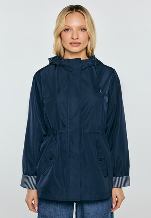 Kobieta ubrana w granatową, wodoodporną kurtkę z kapturem,ściągaczami w paski na mankietach i sznurkiem w talii, stojąca na jednolitym tle.