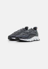 Chaussures de sport grises avec une tige texturée, dotées d'accents noirs et d'une semelle intermédiaire blanche distincte, affichant un design en zigzag.