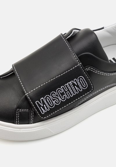 Zapatilla de cuero negro con costuras blancas y una prominente correa de velcro. Cuenta con una suela de goma y un nombre de marca bordado.