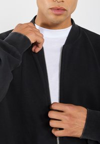 Black bomber jacket with ribbed cuffs, silver zipper, and a relaxed fit. Layered over a white crew-neck shirt. Soft fabric texture.