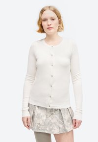 Γυναίκα με κοντά ξανθά μαλλιά που φοράει ένα λευκό ζακέτα με κουμπιά και μια πλισέ γκρι μίνι φλοράλ φούστα σε απλό λευκό φόντο.