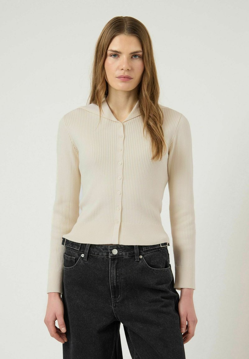 Mujer con cabello largo castaño claro que lleva un cárdigan beige acanalado con botones y jeans negros de talle alto sobre un fondo claro y liso.