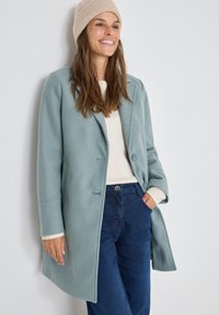 Lichtblauwe wollen jas in een mix van materialen met een gelapt boord, sluiting met één knop en twee zijzakken, gecombineerd met een crèmekleurige trui en blauwe jeans.
