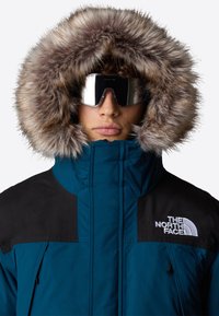 Teal och svart isolerad parka med en huva i päls, som visar en hög krage och en framträdande logotyp på bröstet. Snygga, spegelblanka goggles.