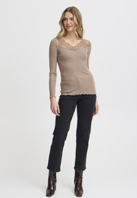Beige Langarmoberteil mit Spitzenausschnitt, strukturiertem Strickstoff und gewelltem Saum, kombiniert mit dunklen Jeans und glänzenden braunen Stiefeletten.