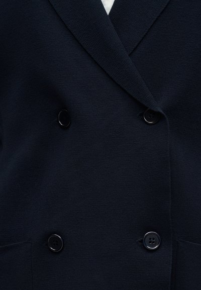 Σκούρο μπλε σακάκι διπλής ραφής με τέσσερα μαύρα κουμπιά και σχισμένους γιακάδες, που δείχνει υφασμένο ύφασμα και μέρος από τις μπροστινές τσέπες.