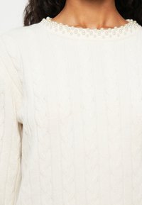 Sweter w warkocze w odcieniu naturalnej bieli, ozdobiony perłami wokół dekoltu; ma prążkowaną fakturę i długie rękawy.