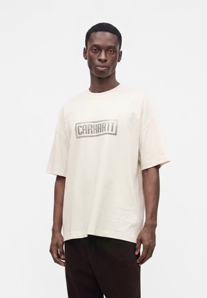 Mężczyzna o neutralnym wyrazie twarzy ubrany w kremowy t-shirt z logo Carhartt i ciemne spodnie na jednolitym białym tle.