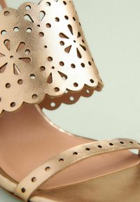 Rose gold sandal with a scalloped strap featuring cut-out floral patterns and small holes. The sole is smooth and has a metallic finish.