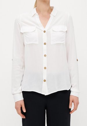 Femme portant un chemisier blanc à manches longues avec des poches avant et des boutons en écaille de tortue, associée à un pantalon noir sur fond blanc.