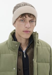Bonnet en tricot beige avec un motif noir, veste puffy verte, et chemise en velours côtelé brun texturé en dessous. Portrait en gros plan sur fond neutre.