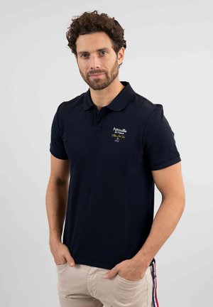 Polo-shirt marine en coton avec un col court, mettant en avant des détails brodés jaunes et blancs. Porté avec un pantalon de couleur claire.